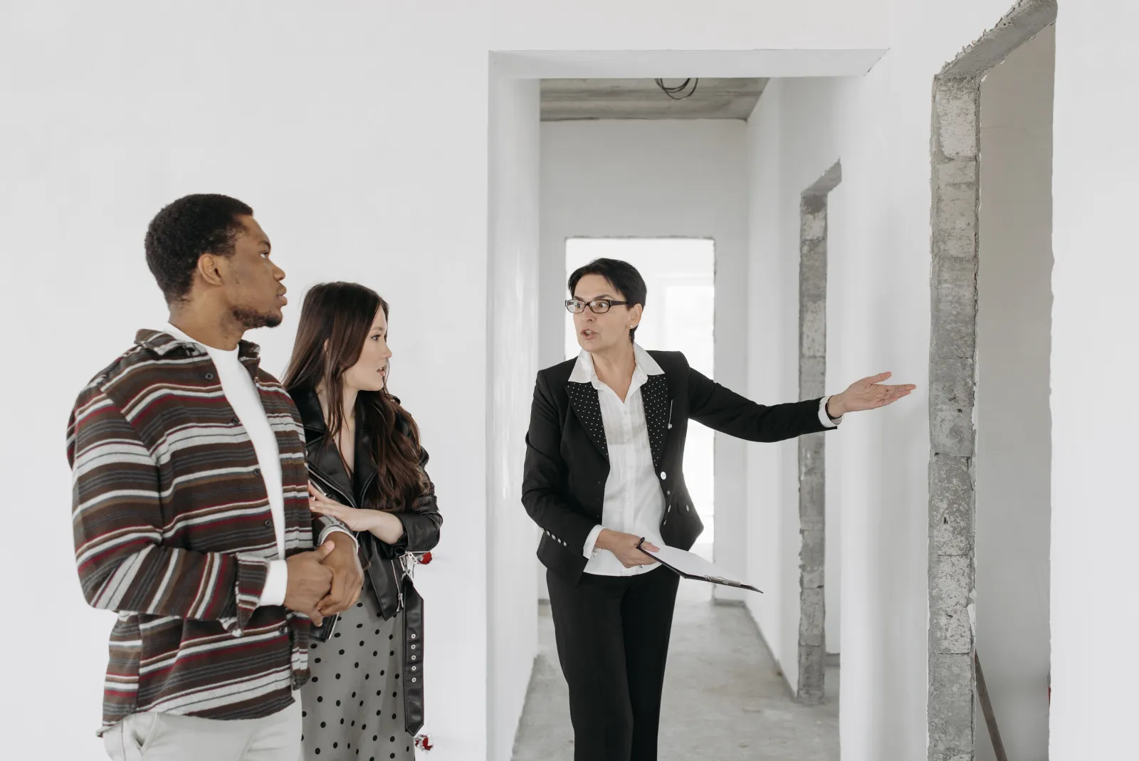 Une professionnelle en tailleur noir, dossier à la main, désigne un mur à un couple lors de la visite d'un logement vide en travaux